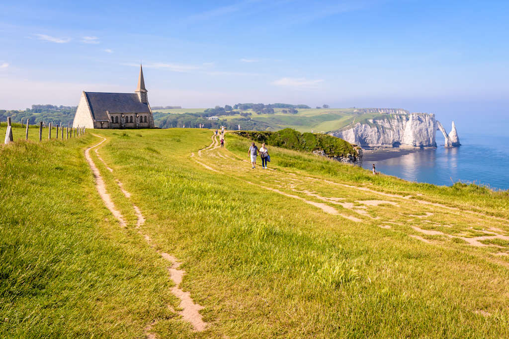 A winding path leads through a lush, grassy field towards a picturesque church, with the dramatic cliffs of the Normandy coast visible in the distance.