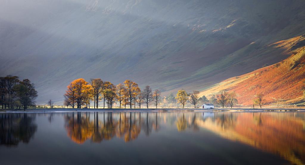 A serene lake surrounded by vibrant autumn foliage, with a mountainous landscape in the background under a dramatic cloudy sky.