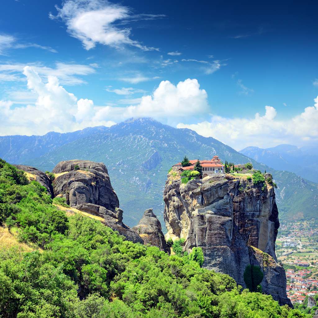 Majestic rock formations rise amidst lush greenery, with a stunning mountain backdrop and a clear blue sky dotted with wispy clouds.