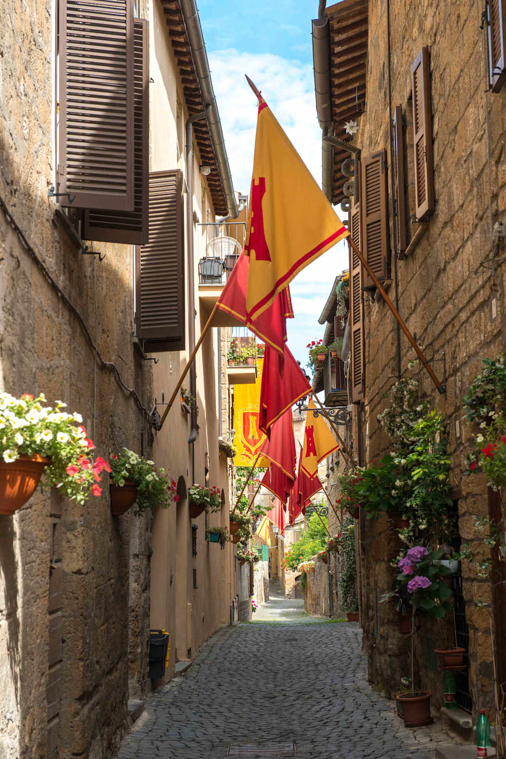 A narrow cobblestone alley lined with colorful flags and potted flowers, surrounded by traditional stone buildings with wooden shutters.