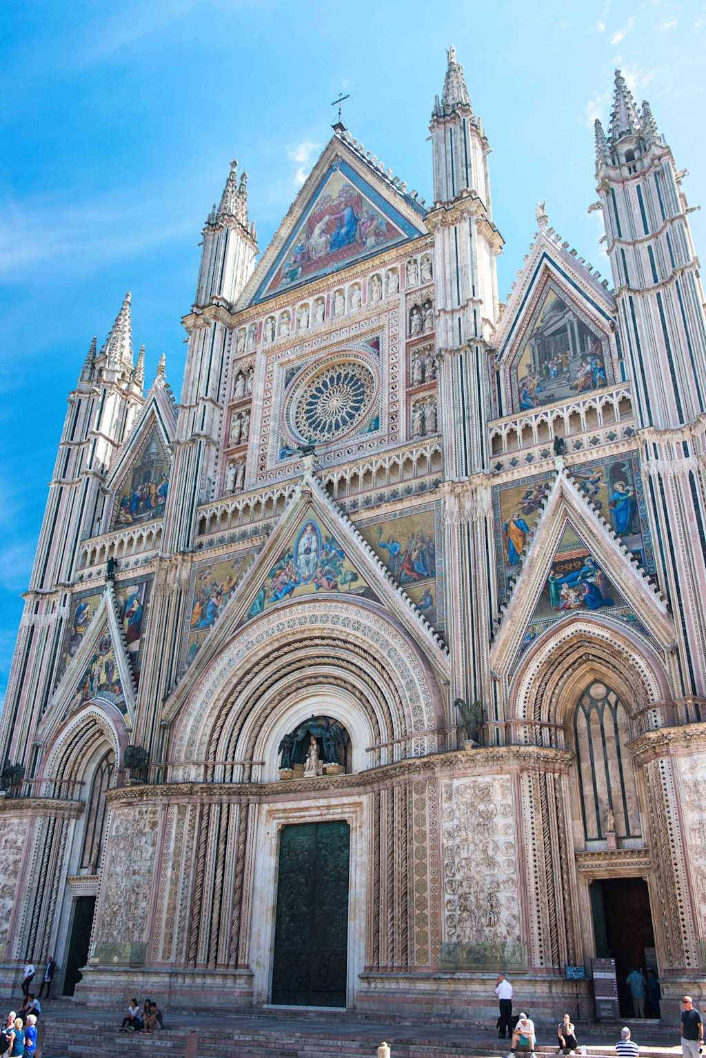 A grand, ornate cathedral with intricate architectural details and decorative elements stands prominently in the foreground, surrounded by a clear blue sky and a few people visible in the immediate vicinity.