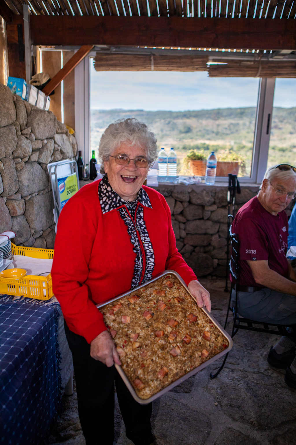 An elderly woman in a red sweater is smiling and holding a baking tray filled with what appears to be a homemade dish, set against the backdrop of a scenic outdoor environment with a stone wall and a view of the landscape beyond.