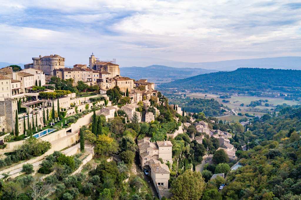 A picturesque medieval town nestled atop a rugged, rocky hillside, surrounded by lush greenery and rolling hills in the distance.