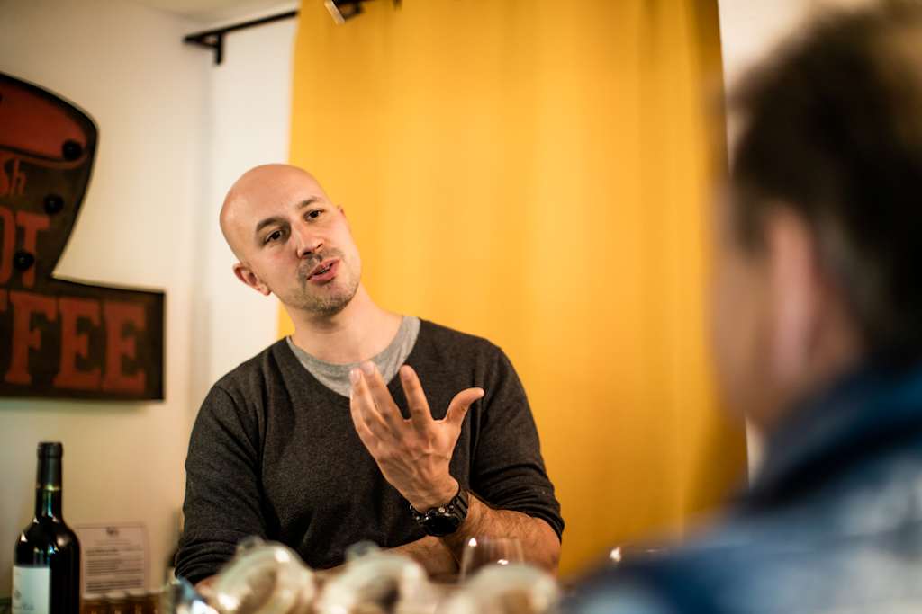 A bald man in a black shirt gesturing while speaking, with a yellow curtain and a framed sign visible in the background.