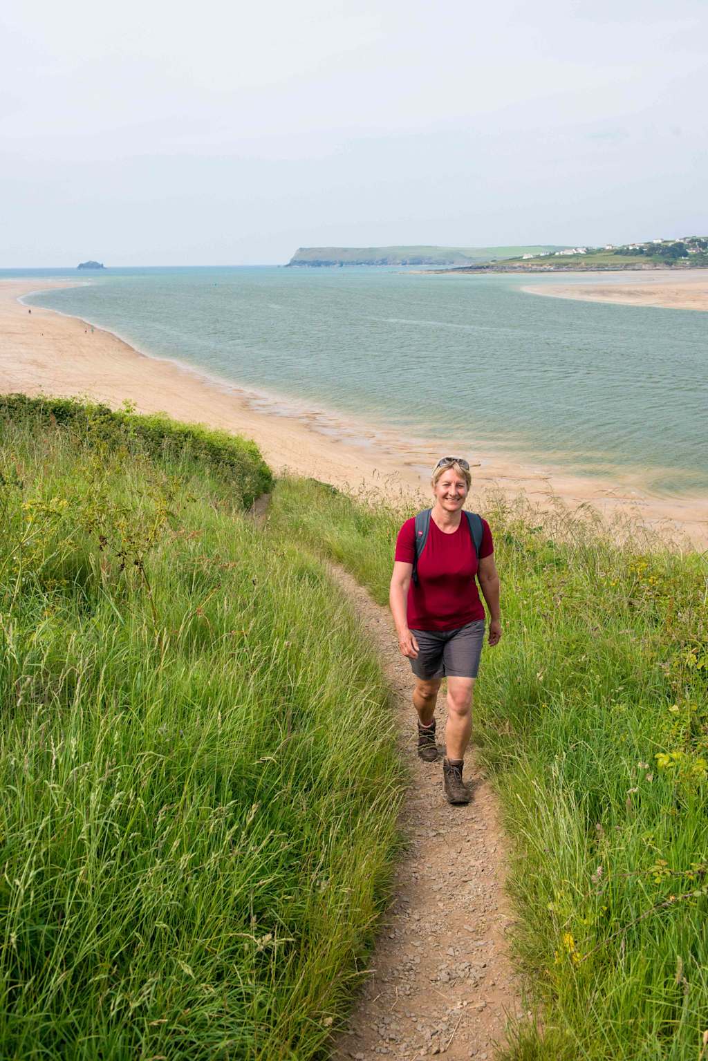 A person in a red shirt and shorts is walking along a dirt path surrounded by lush green grass, with a scenic coastal landscape visible in the background.