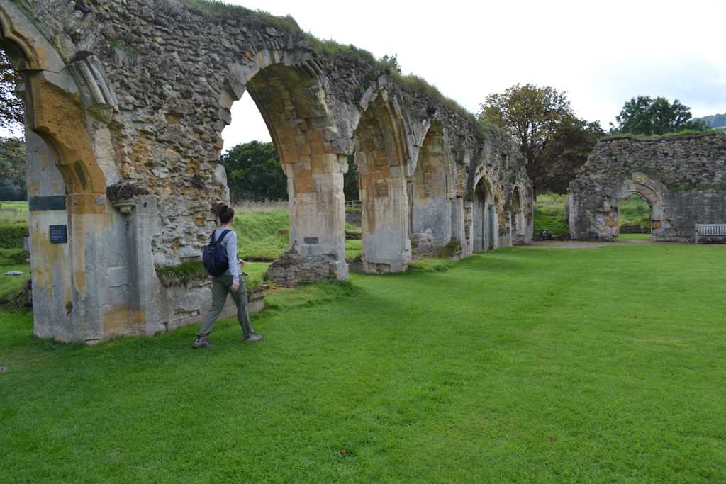 The image depicts a series of stone arches surrounded by lush greenery, with a person walking along the grassy path in the foreground.