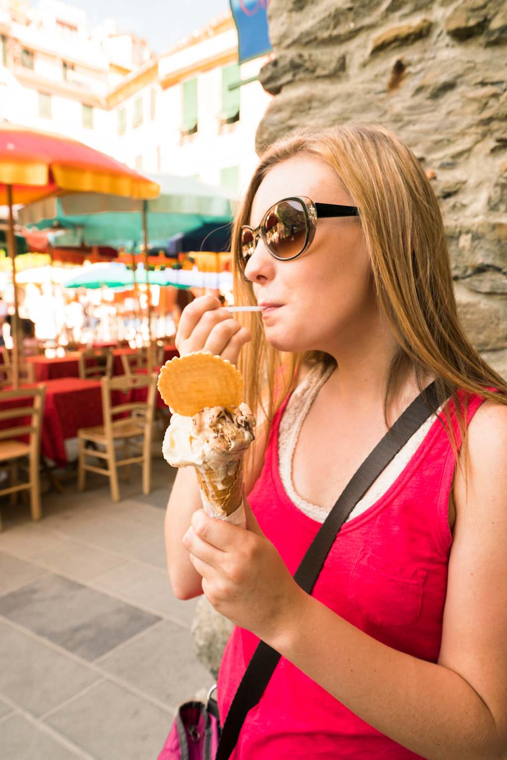 A woman in a red top and sunglasses is enjoying an ice cream cone while sitting at an outdoor dining area with colorful umbrellas and tables.