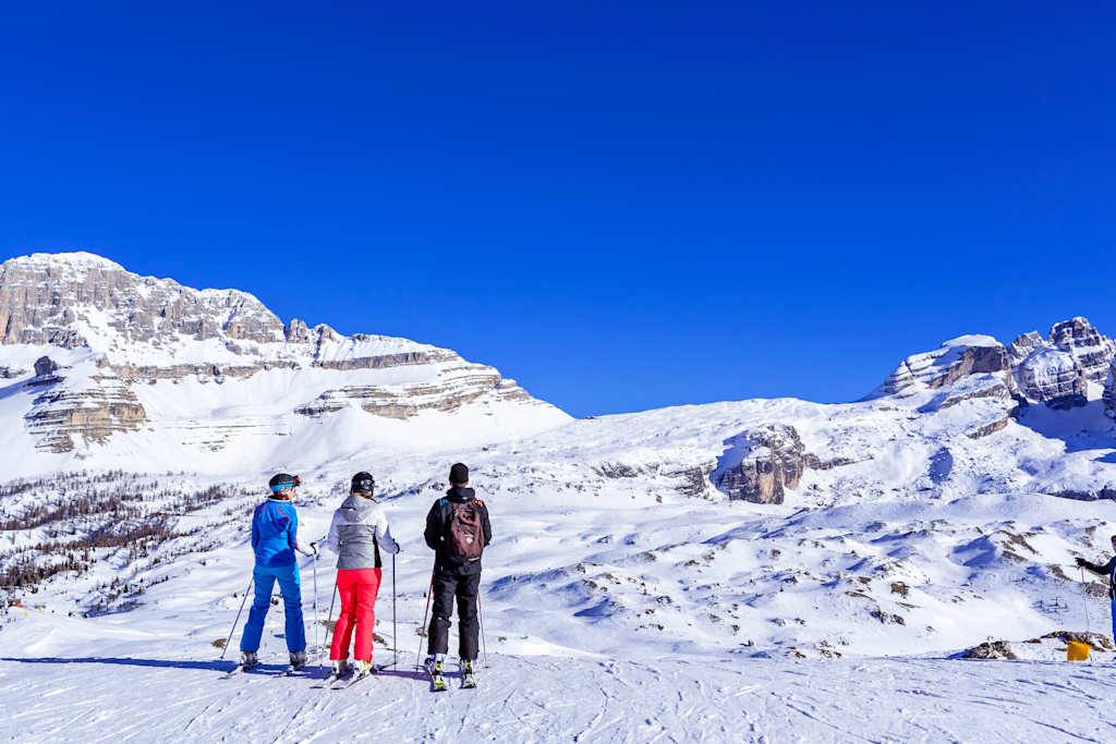 Three people standing on a snowy mountain slope, with a stunning backdrop of snow-capped peaks against a vibrant blue sky.