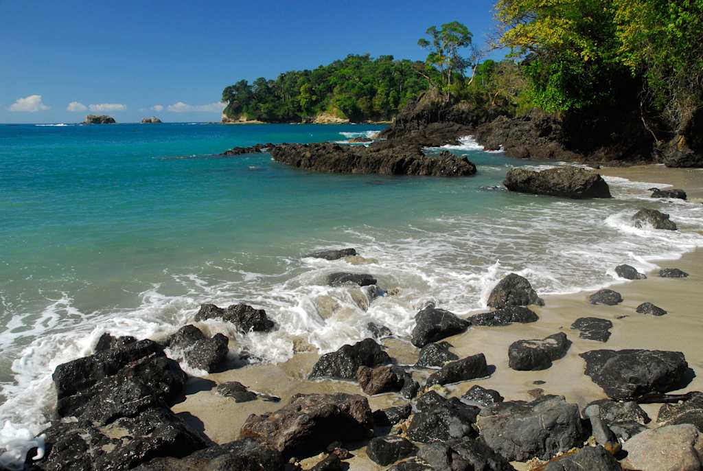 A picturesque tropical beach with crystal-clear turquoise waters, rocky outcroppings, and lush green foliage in the background.