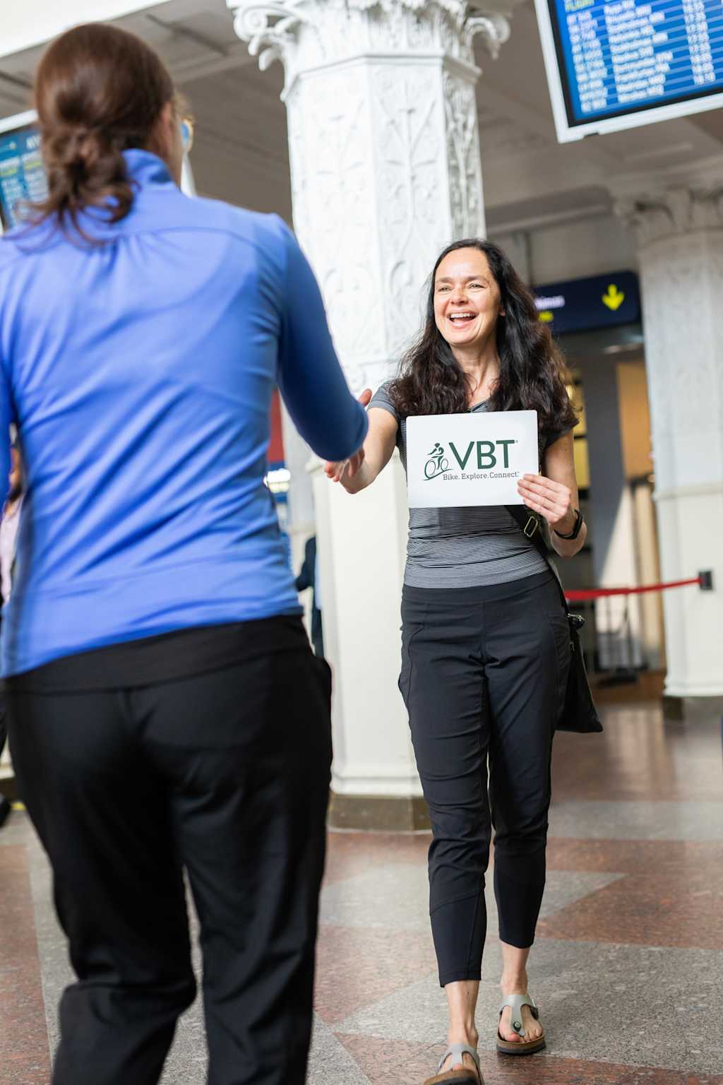 The image shows a smiling woman holding a sign that says "VBT" in an airport terminal, with a person in the background.