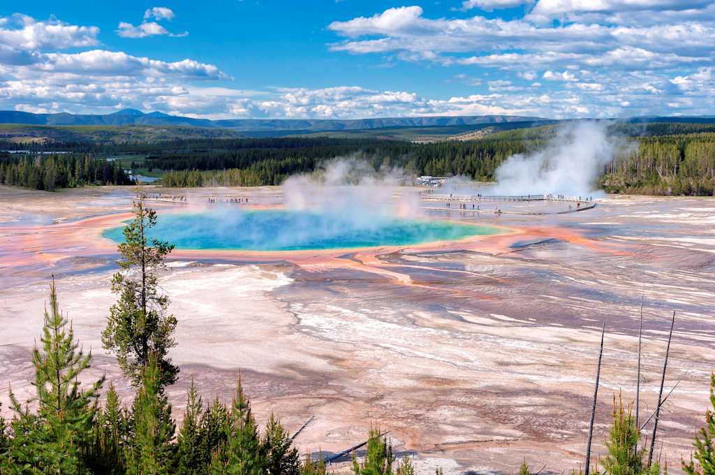 A stunning natural landscape featuring a vibrant, multi-colored hot spring surrounded by a rugged, forested terrain under a vast, cloudy blue sky.