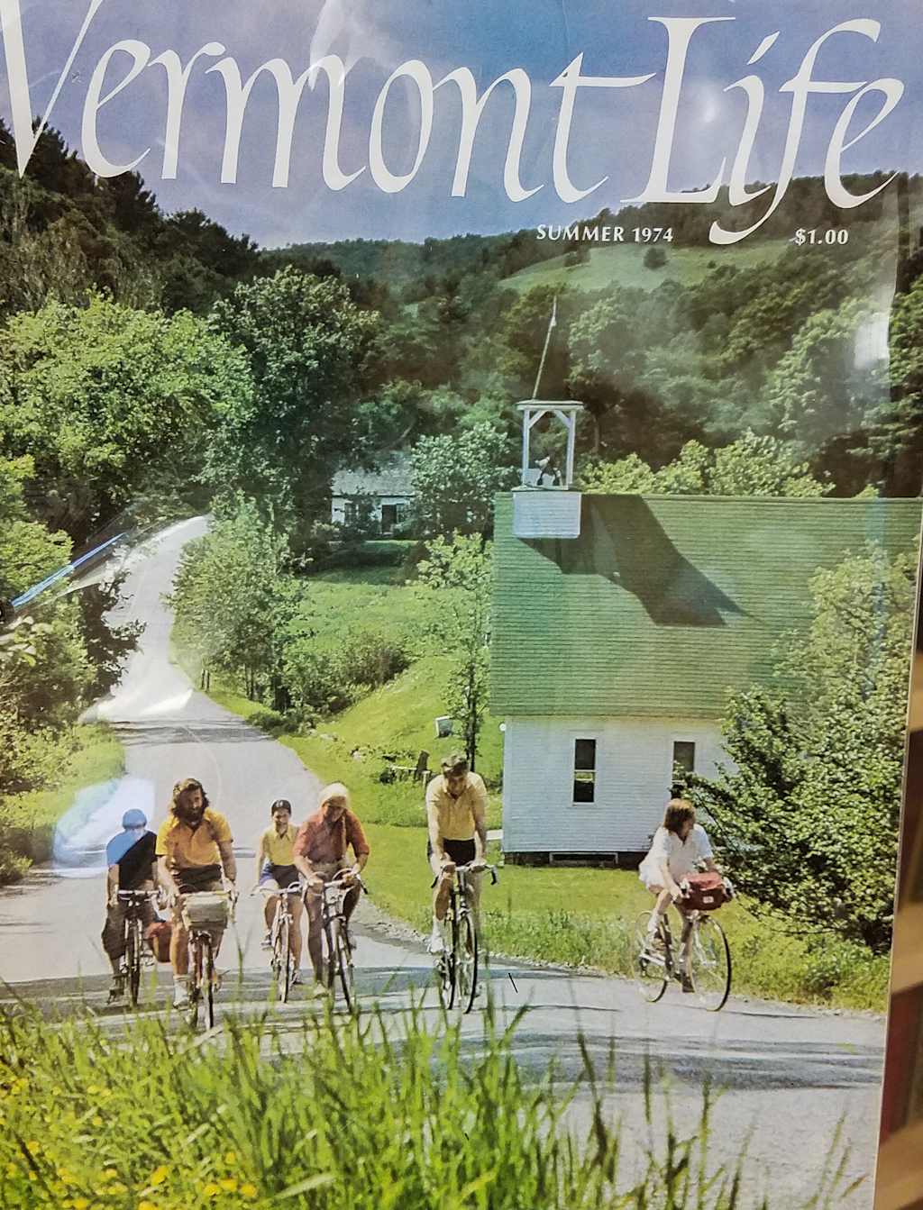 A group of people riding bicycles on a rural road surrounded by lush greenery and a picturesque Vermont landscape in the background.