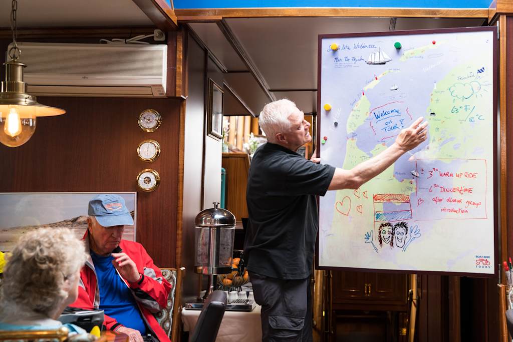 An older man in a black shirt is standing in front of a large whiteboard, gesturing and writing on it, while several people are seated in the background.