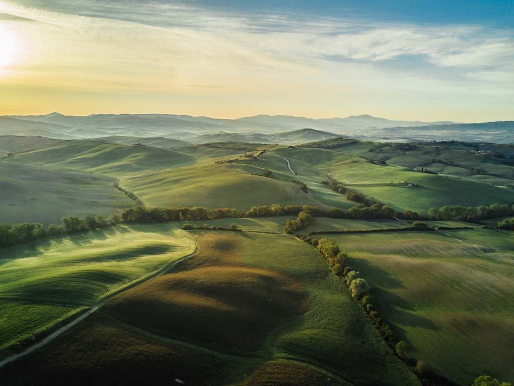 A picturesque landscape with rolling green hills, winding roads, and distant mountains bathed in a warm, hazy light, creating a serene and tranquil atmosphere.