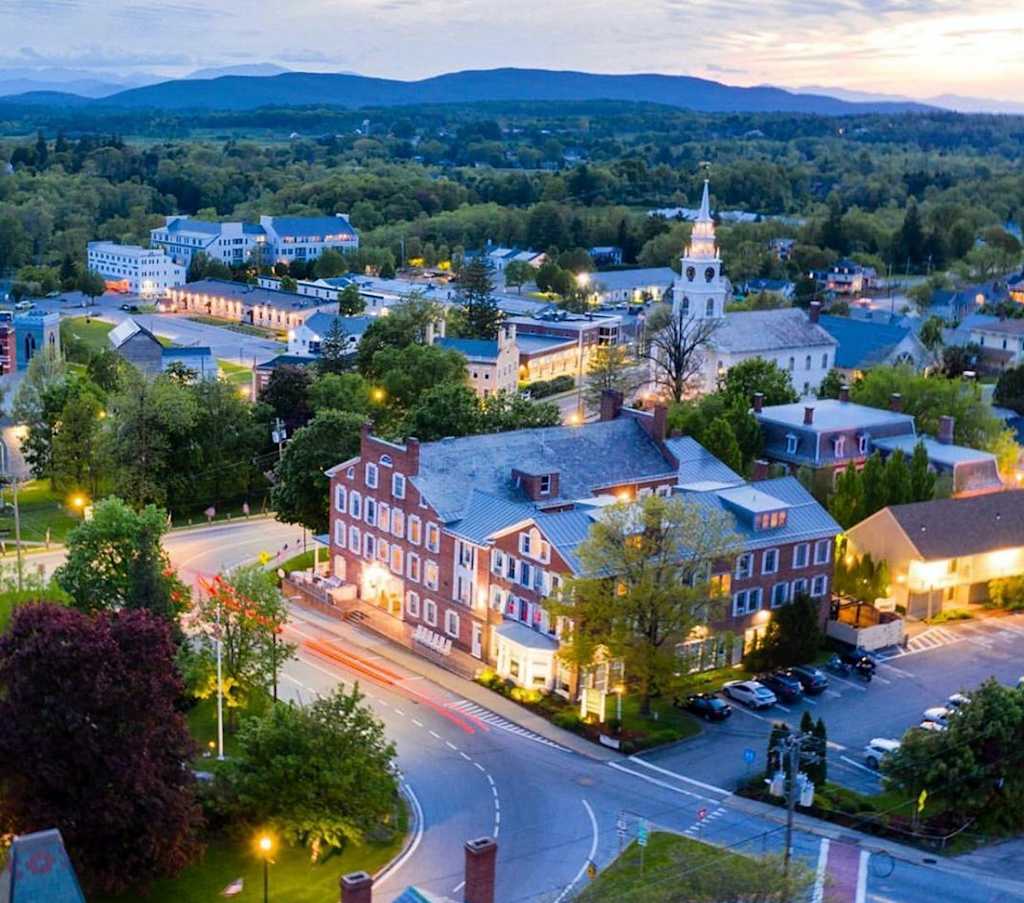 A picturesque town nestled in a valley, with a church steeple rising above the colorful buildings and streets illuminated by the warm glow of evening lights, surrounded by lush greenery and distant mountains.