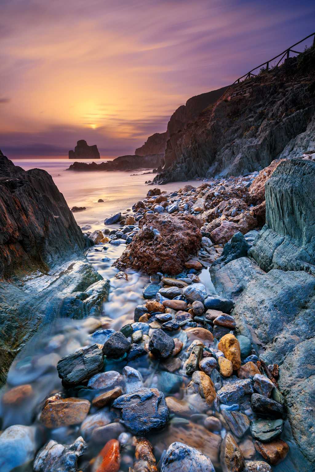 A breathtaking coastal landscape at sunset, with a rocky foreground leading to a serene body of water and a vibrant sky filled with warm hues.