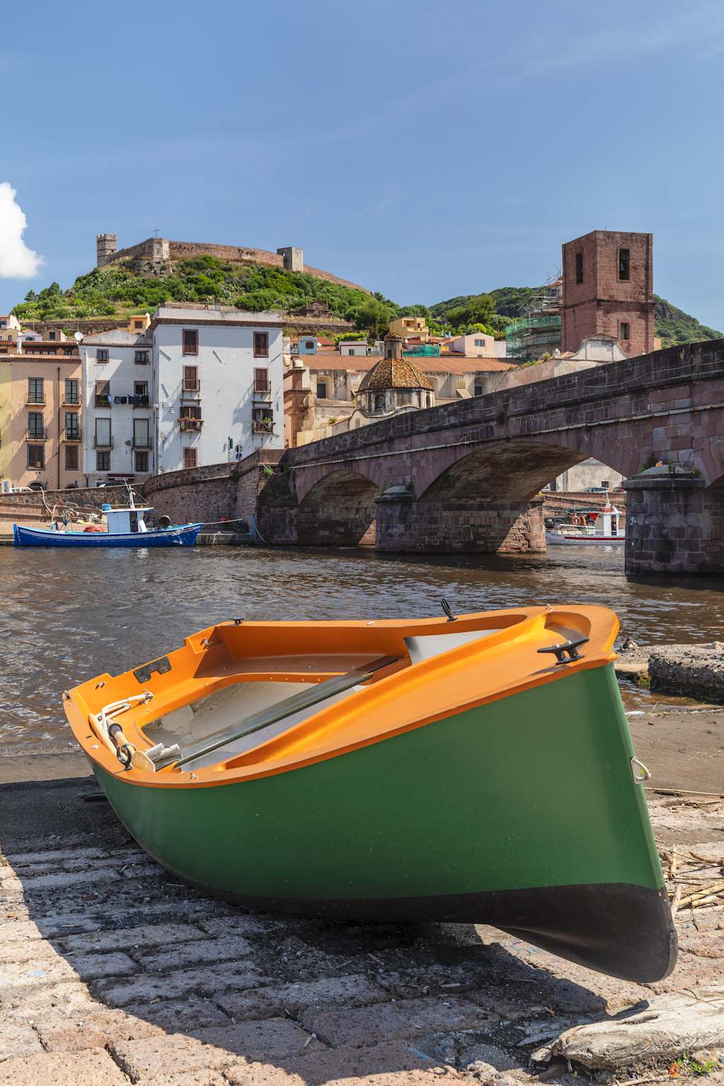 A small green and orange boat rests on the shore, with a historic town and castle visible in the background against a clear blue sky.
