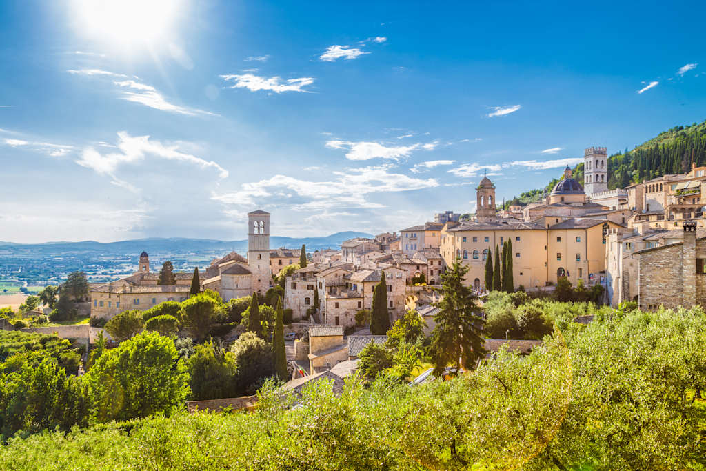A picturesque hillside town with colorful buildings, churches, and towers nestled among lush green trees, set against a bright blue sky with fluffy white clouds.