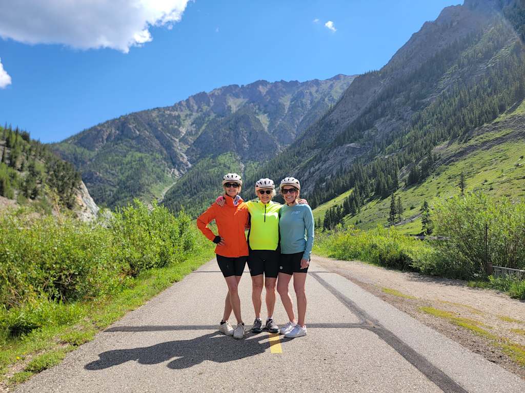 Three individuals in athletic gear stand on a dirt road surrounded by lush green vegetation and towering mountains in the background.