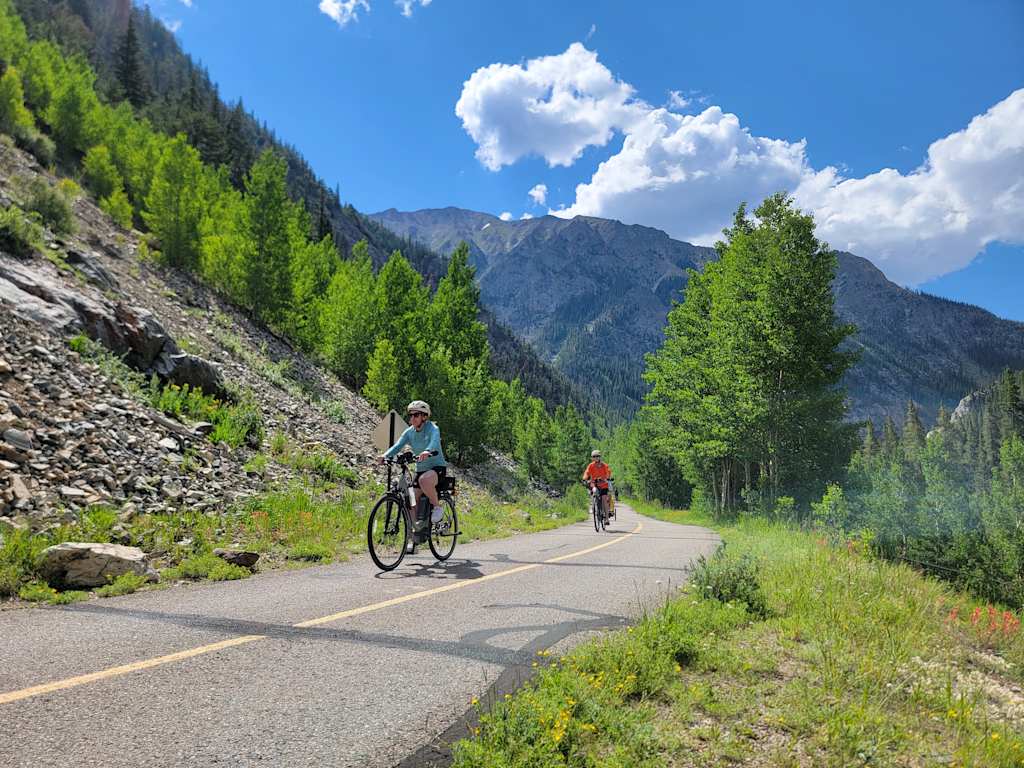 A scenic mountain road with cyclists riding through a lush, forested landscape under a blue sky with fluffy clouds.