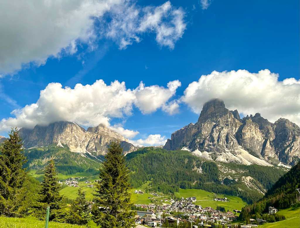 Majestic snow-capped mountains rise against a vibrant blue sky dotted with fluffy white clouds, while a quaint village nestles in the lush green valley below, surrounded by towering evergreen trees.