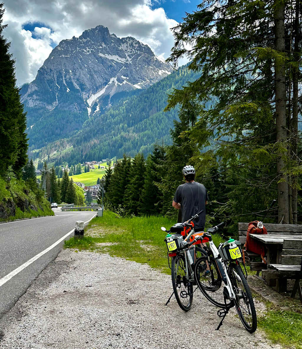 A cyclist stands on a road surrounded by lush green forests and towering snow-capped mountains in the distance.