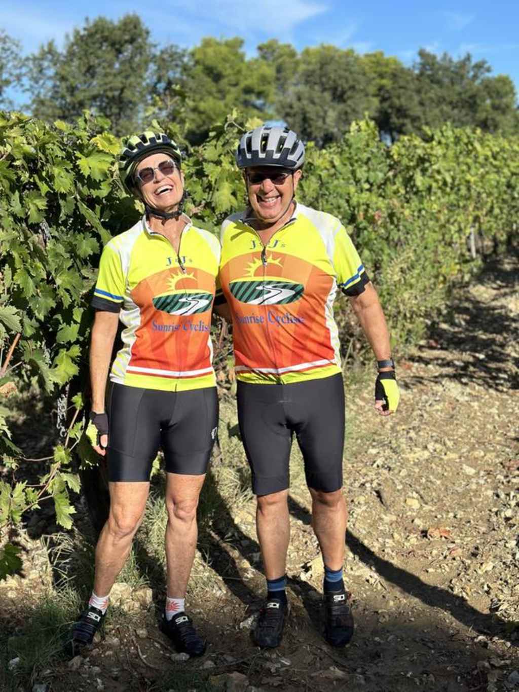 Two cyclists in bright cycling gear stand on a dirt path surrounded by lush greenery and trees.