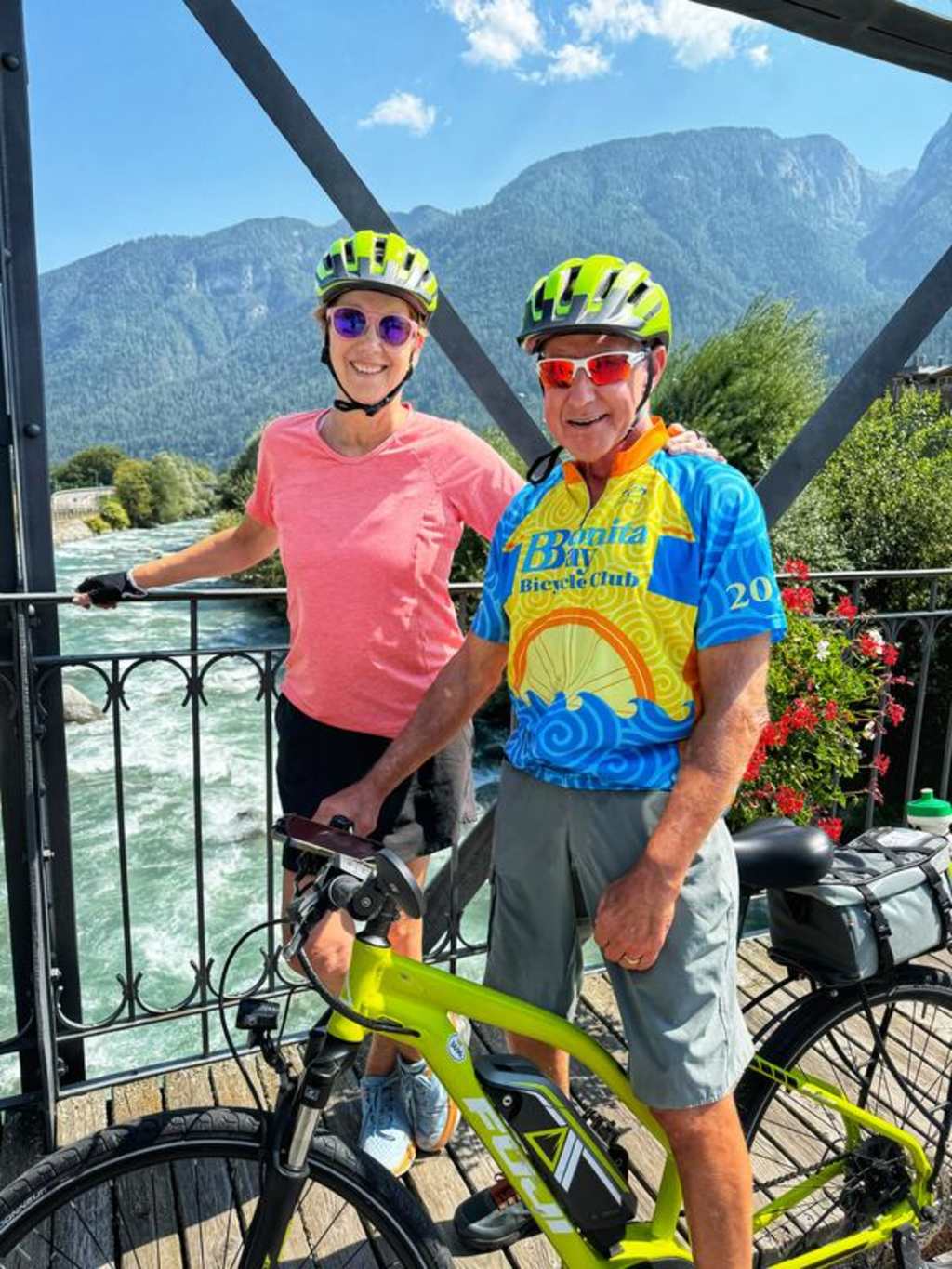 Two cyclists, a man and a woman, are posing on a balcony with a scenic mountain landscape in the background.