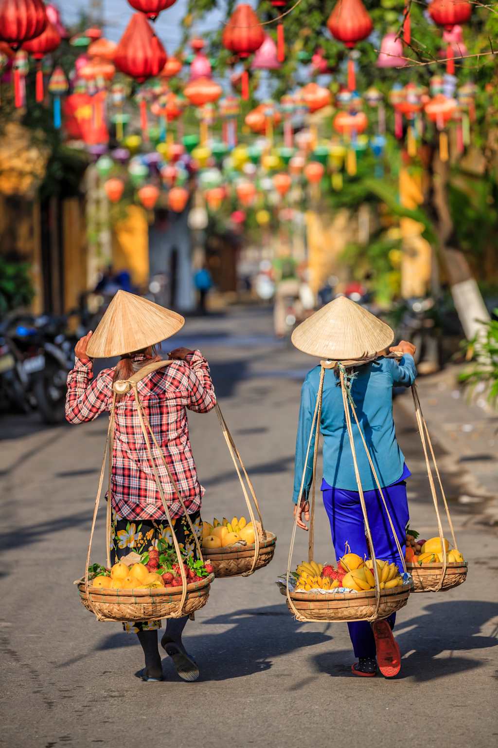 Two people wearing traditional conical hats and carrying baskets of fruits and vegetables on a street lined with colorful lanterns and buildings.