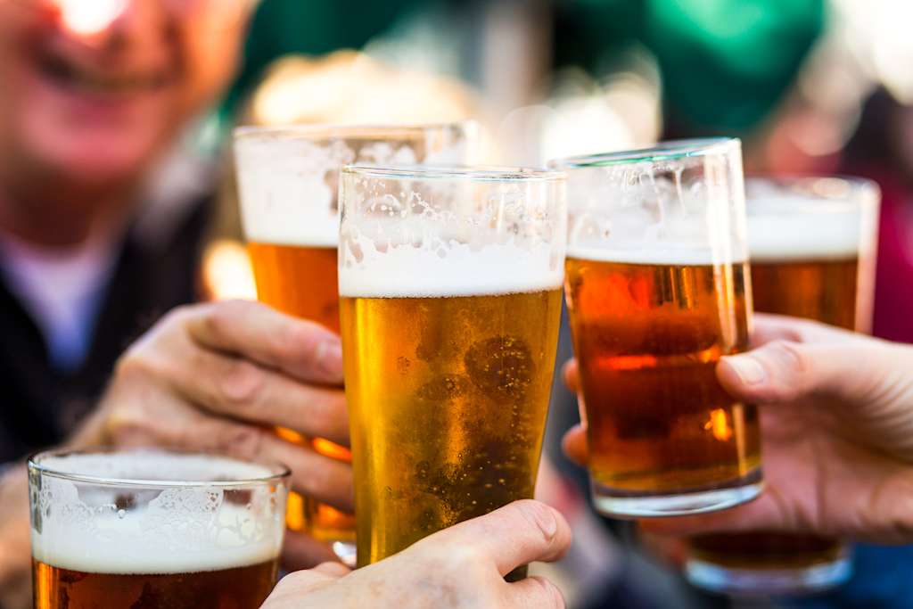The image shows several glasses of beer being held by people's hands, with a blurred background suggesting a social gathering or bar setting.