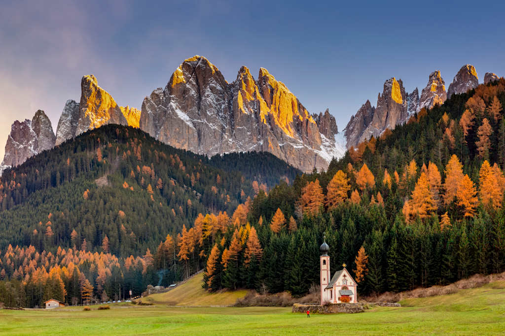 A picturesque landscape featuring towering, jagged mountains in the background, surrounded by a vibrant display of autumn foliage, with a small church nestled in the foreground against a lush, grassy meadow.