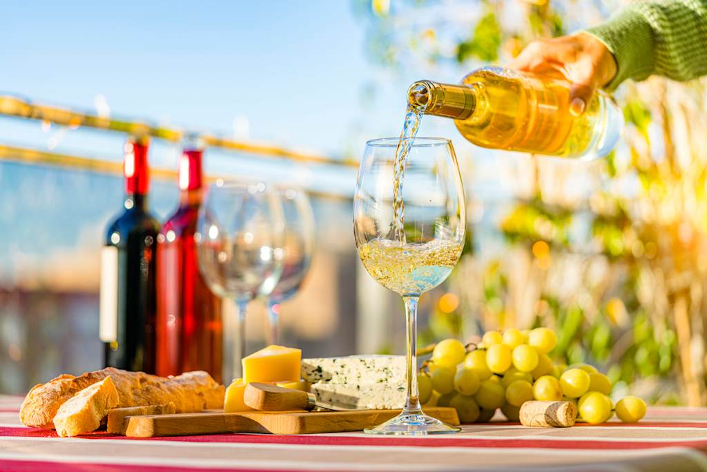 A glass of white wine being poured, surrounded by various cheeses, grapes, and a bottle of red wine, set against a backdrop of a sunny outdoor scene with greenery.