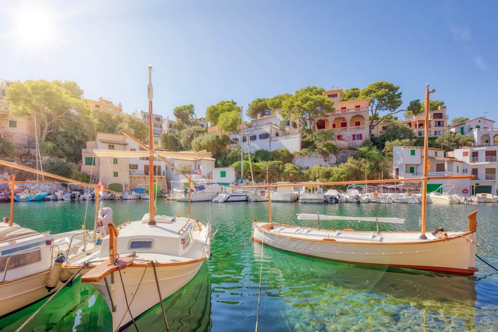 A picturesque harbor with colorful boats moored in the calm, turquoise waters, surrounded by charming Mediterranean-style buildings nestled among lush greenery under a bright, clear sky.