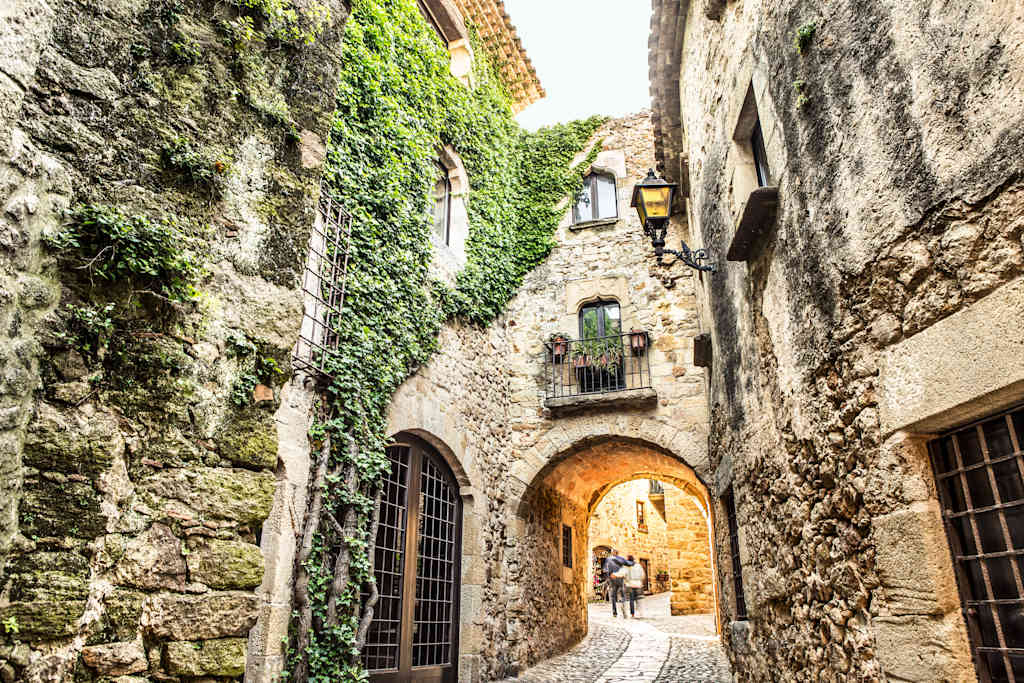 A narrow, winding alleyway lined with stone walls and covered in lush greenery, leading to a sunlit archway in the distance.
