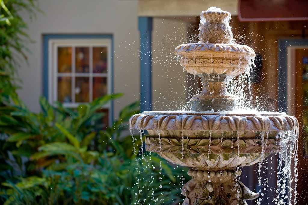 A decorative stone fountain with cascading water stands in a lush, verdant garden setting, surrounded by potted plants and a building with a blue-framed window in the background.