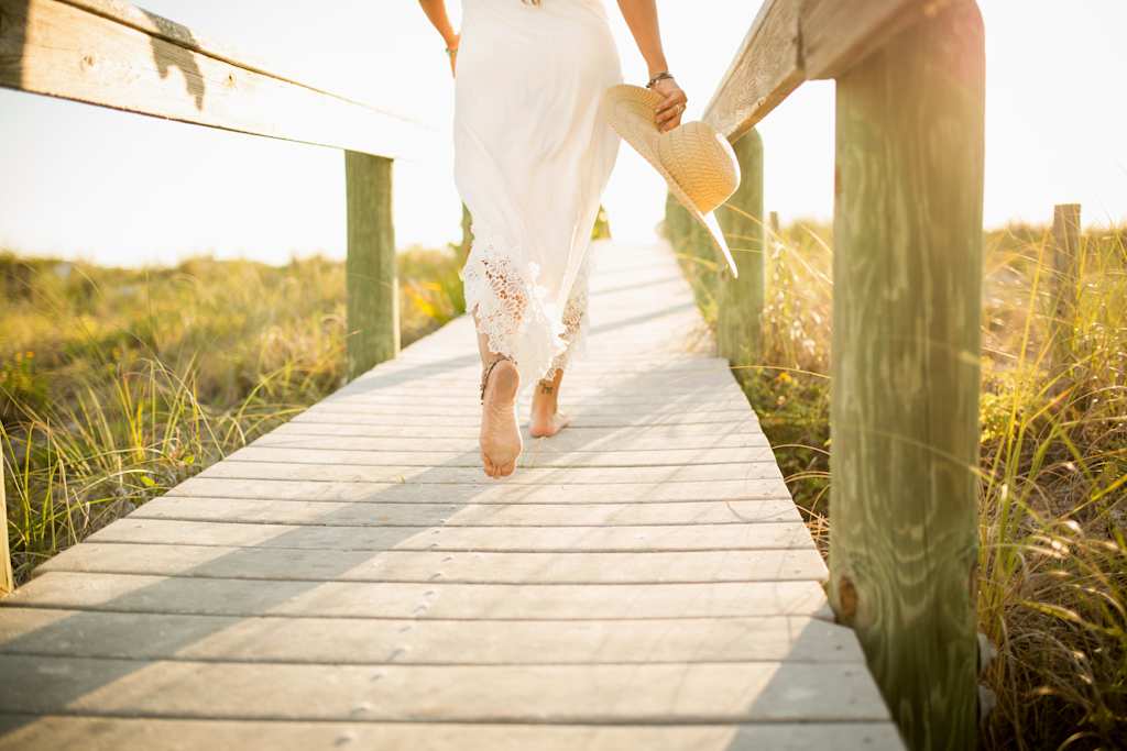 A person in a white dress walks barefoot along a wooden boardwalk surrounded by tall grass and a scenic countryside landscape.