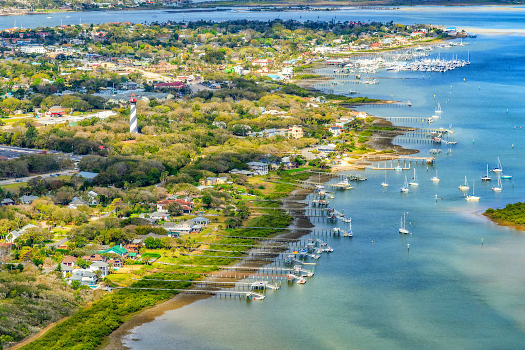 A picturesque coastal town nestled along a serene body of water, with a mix of residential and commercial buildings, a marina filled with boats, and lush greenery surrounding the area.