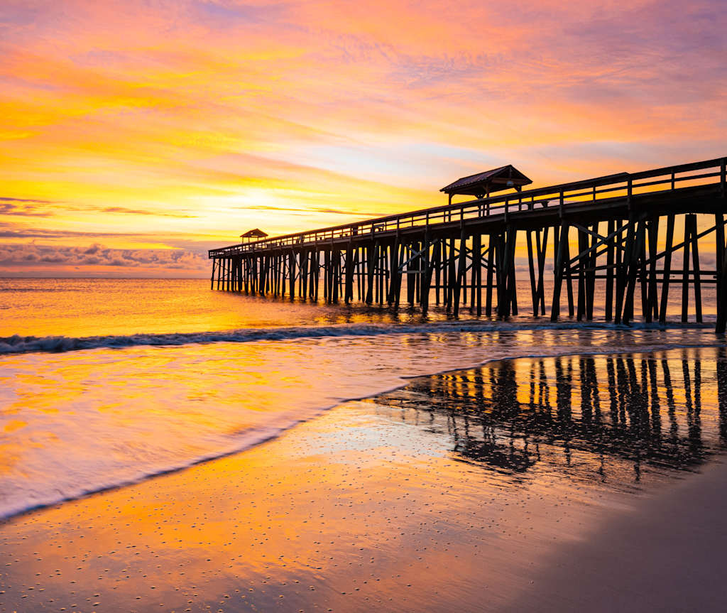 A wooden pier extends into a tranquil ocean, reflecting the vibrant orange and yellow hues of the stunning sunset in the background.