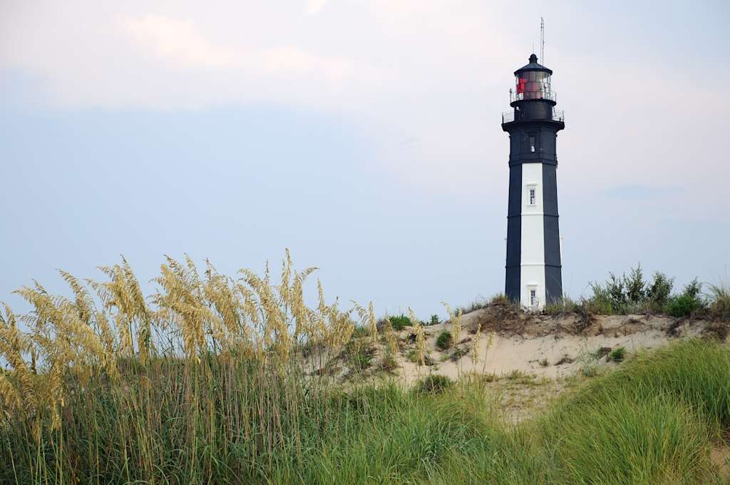 A tall, black-and-white lighthouse stands prominently against the sky, surrounded by tall grass and sand dunes in the foreground.