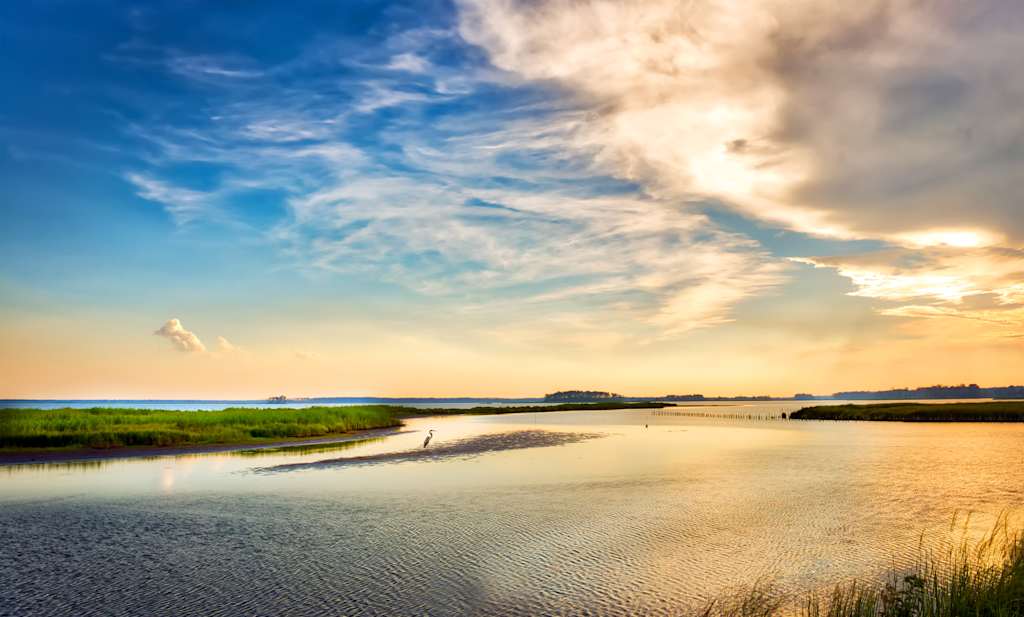 A serene coastal landscape with a tranquil waterway surrounded by lush marshlands, set against a vibrant sky with dramatic clouds at sunset.