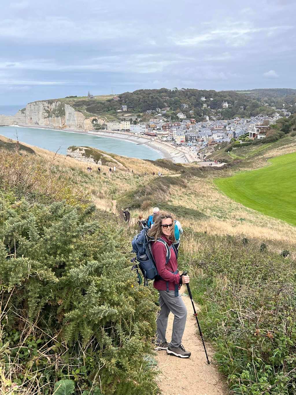 A hiker with a backpack walks along a trail overlooking a coastal town nestled between hills and the ocean.