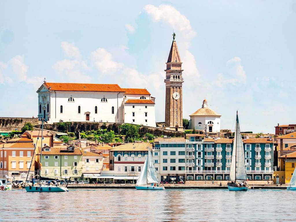 A picturesque coastal town with colorful buildings, a tall church steeple, and boats docked along the waterfront, set against a backdrop of a bright, cloudy sky.