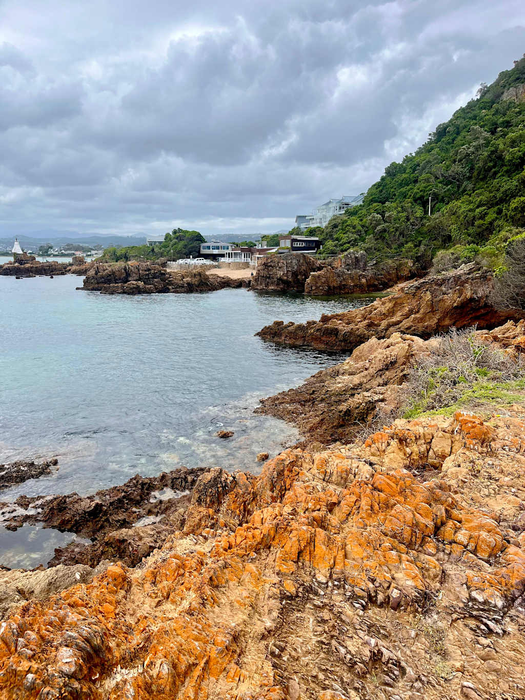 A rugged, rocky coastline with a calm, turquoise sea in the foreground, surrounded by lush, green hills and a cloudy, overcast sky in the background.