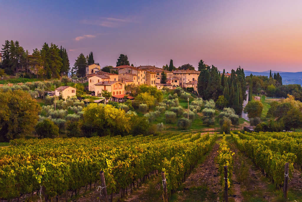 A picturesque Italian village nestled among rolling vineyards, with a stunning sunset backdrop illuminating the charming architecture and lush greenery.