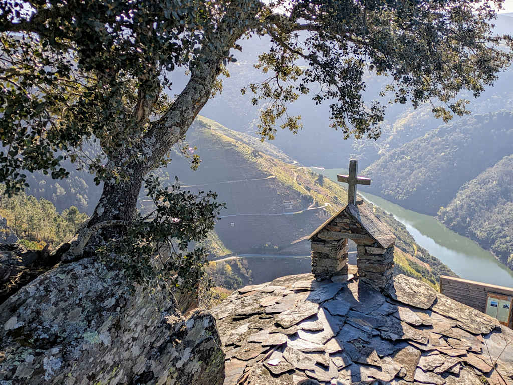 A stone chapel with a cross atop sits on a rocky outcrop, overlooking a serene lake surrounded by lush, forested mountains in the distance.