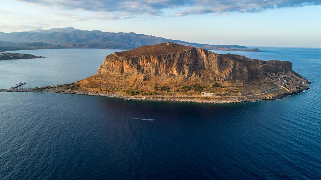 A rugged, rocky peninsula juts out into a vast, deep blue ocean, surrounded by towering mountains and a dramatic, cloudy sky.
