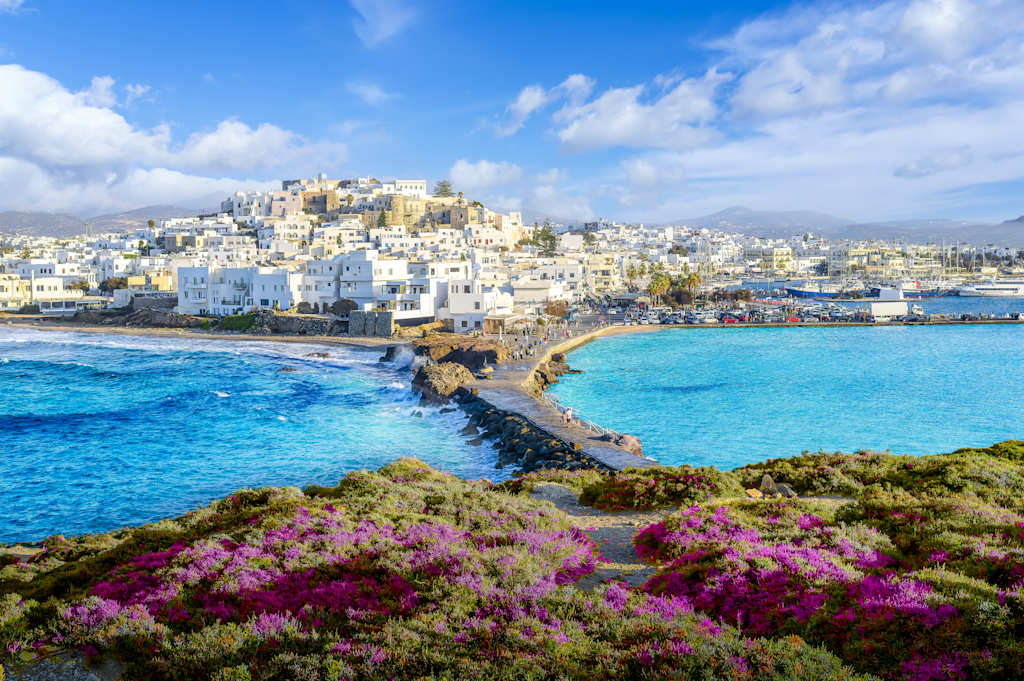 A picturesque coastal town with white buildings nestled among vibrant purple flowers in the foreground, overlooking a stunning turquoise sea under a bright blue sky with fluffy clouds.