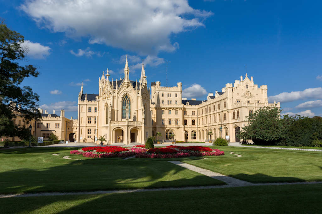 A grand, ornate castle-like structure with multiple towers and spires stands amidst a well-manicured lawn and garden, set against a backdrop of a bright blue sky with fluffy white clouds.
