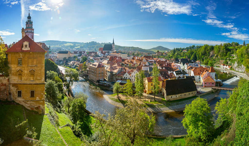 A picturesque medieval town nestled in a lush, verdant valley, with a winding river and historic buildings adorning the landscape under a vibrant blue sky.