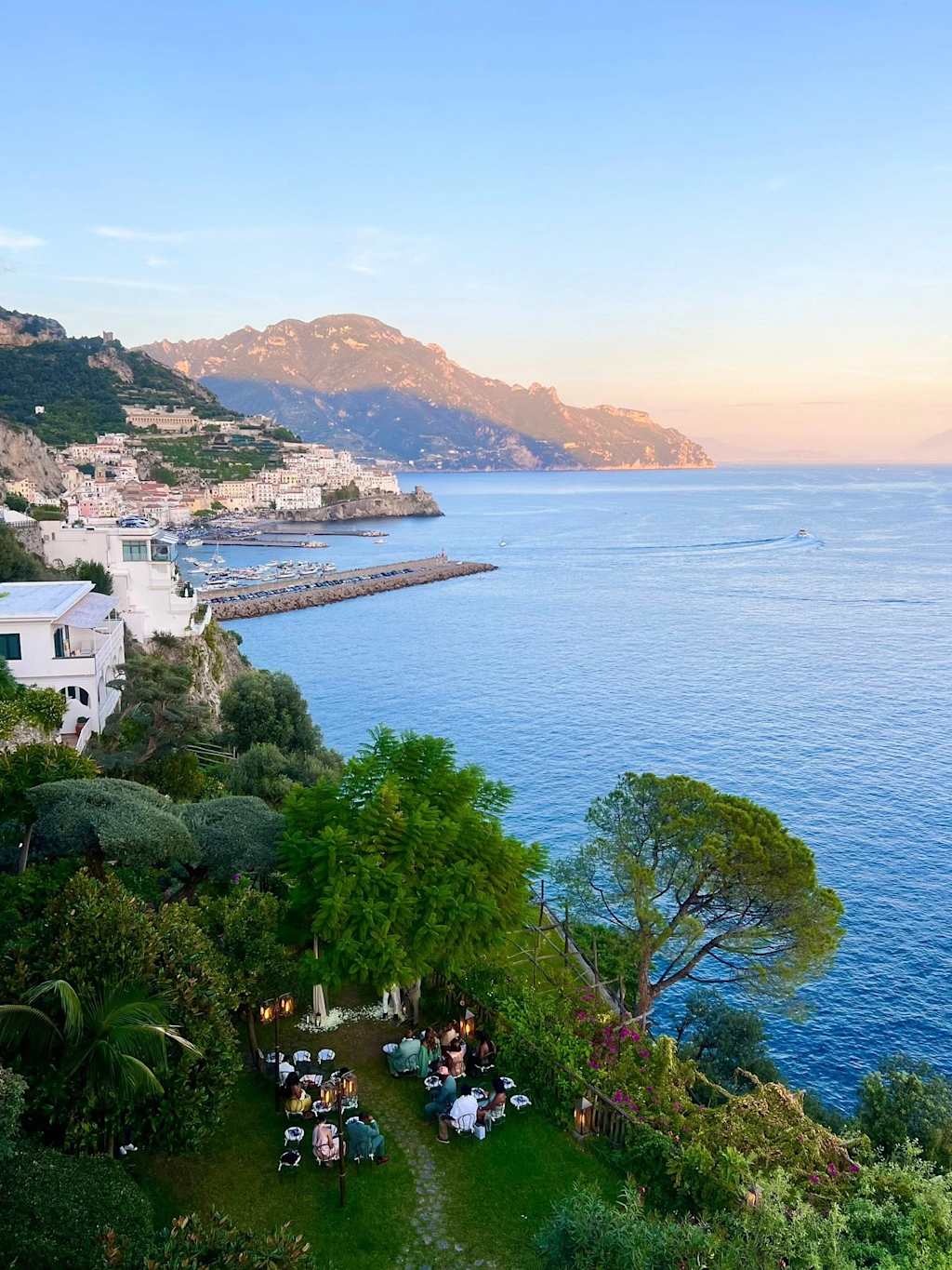 A picturesque coastal town nestled among lush greenery, with a scenic harbor and distant mountains visible under a clear blue sky.